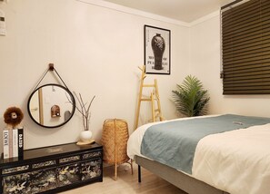 Ein Schlafzimmer mit einem Bett, einer Kommode mit Dekorationen, einem Spiegel, einem gerahmten Bild an der Wand, einer Hockerleiter und einer Topfpflanze.