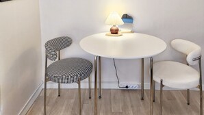 Et lille, rundt bord med en lampe og en pynteting hængende over. To stole med et hundetandsmønster og en stol med en fluffy sæde. Væggen er hvid og gulvet er af træ.