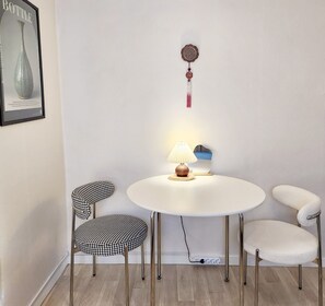 Ein kleiner, runder Tisch mit einer Lampe und einem dekorativen Gegenstand darüber. Es gibt zwei Stühle mit Hundszahn-Muster und einen mit Sitzfläche aus Flauschmaterial. Der Raum hat eine weiße Wand und Holzdielen.