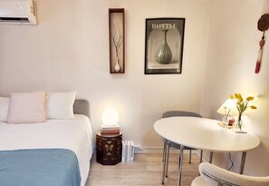 Quarto de hotel com cama, mesa de cabeceira com abajur, uma obra de arte emoldurada, uma pequena mesa com cadeira e um objeto decorativo pendurado.