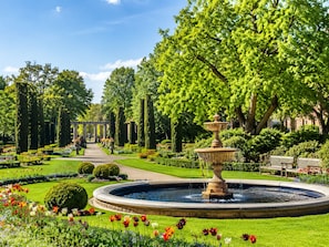 Ein formeller Garten mit einem zentralen Brunnen, ordentlich geschnittenen Hecken und einer Vielzahl von Blumen.