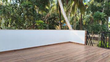 A wooden deck with a white wall and a metal railing, surrounded by lush greenery.