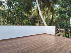 Una terraza de madera con un muro blanco y barandilla metálica, rodeada de abundante vegetación.