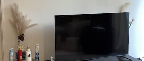 En stue med et fladskærms TV på et sort stativ, et lille bord med rengøringsmidler og et vægur.