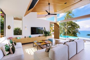Un soggiorno moderno con una grande televisione, posti a sedere comodi, un soffitto in legno e vista sull'oceano.