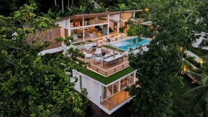 Een modern huis met twee verdiepingen, een dakterraszwembad en een buiten zitgedeelte omgeven door weelderige begroeiing.