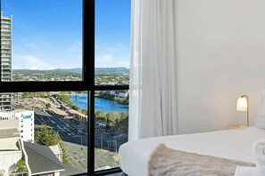 Ein Hotelzimmer mit großem Fenster und Blick auf die Stadt, einem Bett mit weißer Bettdecke und einer Nachttischlampe.