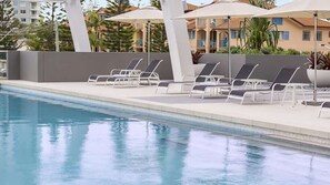 En swimmingpool med liggestole og parasoller, omgivet af en lav mur og bygninger i baggrunden.