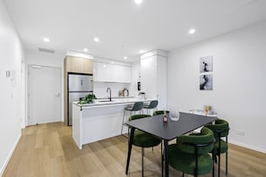A modern kitchen with white cabinets, a central island, and a dining area with a table and green chairs. - Riverfront 2BR Surfers Paradise Apt, Pool, Gym & Free Parking (Surfers Paradise)
