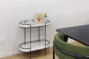 Een eethoek met een zwarte tafel en een groene stoel, een metalen plank met een fotolijstje en een fles, en een marmeren aanrecht.