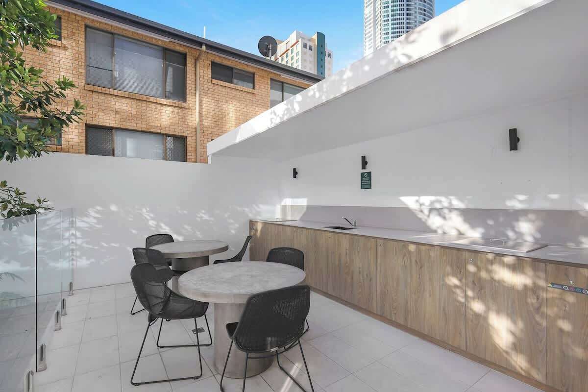 A modern outdoor seating area with a round table and chairs, a wooden counter with a sink, and a glass partition.