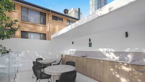 A modern outdoor seating area with a round table and chairs, a wooden counter with a sink, and a glass partition. - Riverfront 2BR | Pool + Gym | Surfers (Surfers Paradise)