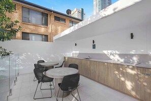 A modern outdoor seating area with a round table and chairs, a wooden counter with a sink, and a glass partition. - Riverfront 2BR | Pool + Gym | Surfers (Surfers Paradise)