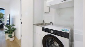 A modern laundry room with a front-loading washing machine, a sink, and a potted plant. - Riverfront 2BR | Pool + Gym | Surfers (Surfers Paradise)