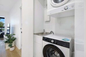 A modern laundry room with a front-loading washing machine, a sink, and a potted plant. - Riverfront 2BR | Pool + Gym | Surfers (Surfers Paradise)