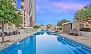 A swimming pool with lounge chairs, surrounded by tall buildings and greenery. - 2BR Ocean View Hilton | Kids' Pool, Gym, Parking (Surfers Paradise)