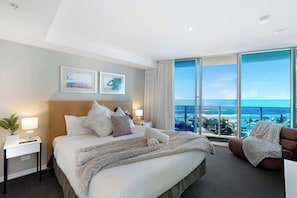 Ein Hotelzimmer mit einem großen Bett, zwei Nachttischen, einem Sessel und einem großen Fenster mit Blick aufs Meer.