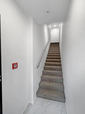 Una escalera con barandilla, un detector de incendios en la pared y una lámpara en el techo.