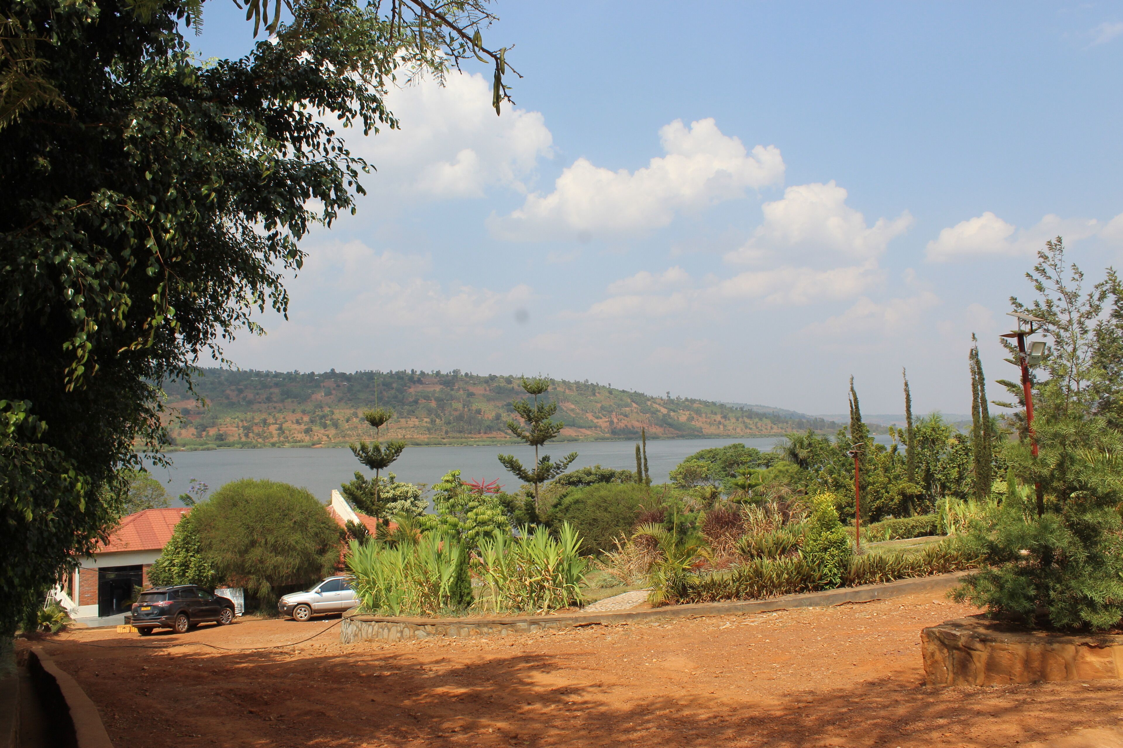 A view of a lake with a hilly landscape in the background, surrounded by lush greenery and a few parked cars.