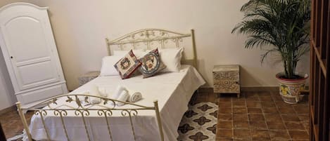 Una cama bien tendida con sábanas blancas y almohadas decorativas, una mesita de noche con lámpara, y una planta en maceta en la esquina.