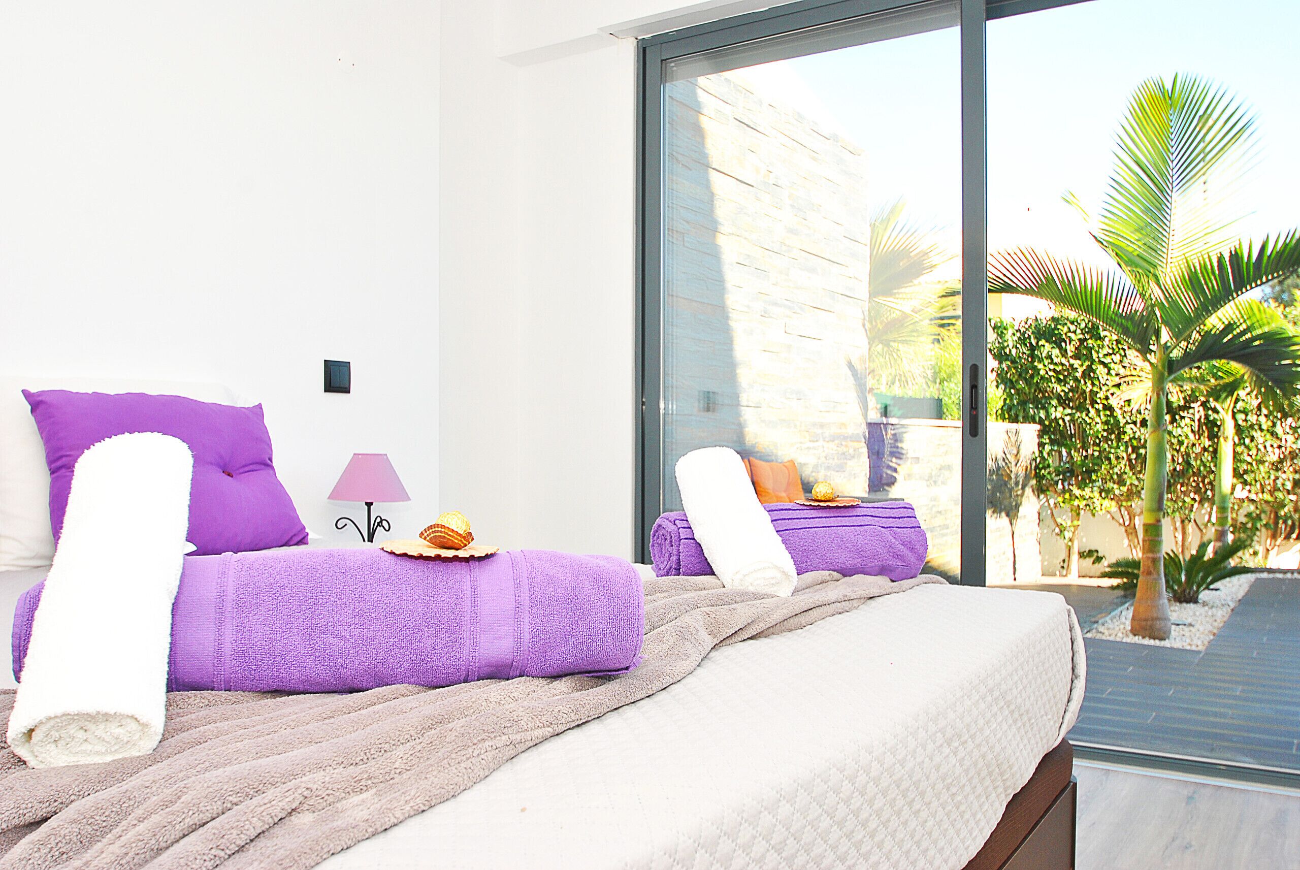 Un dormitorio con un ventanal grande, una cama con ropa de cama morada, una lámpara y una mesita con un objeto decorativo.