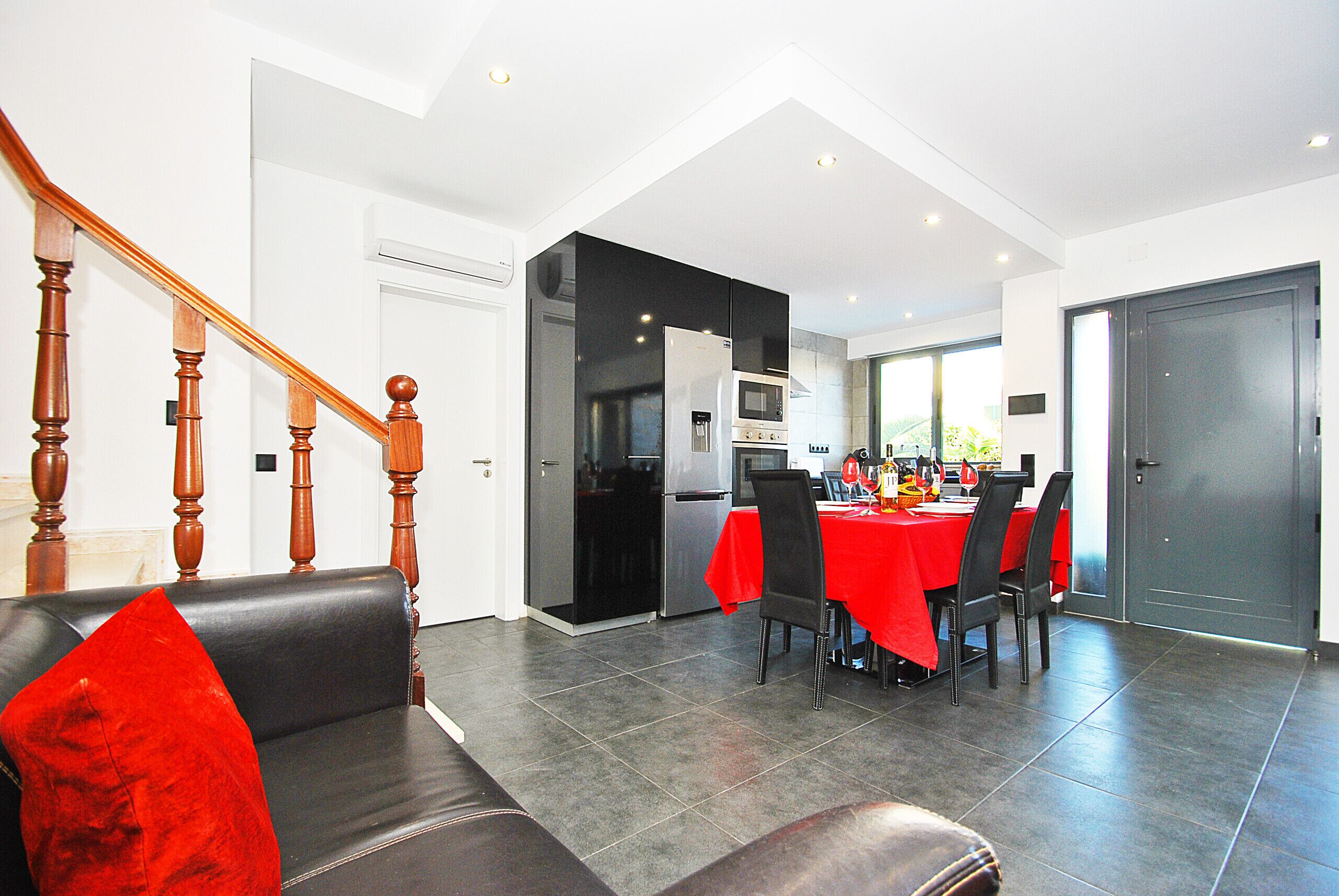 Una cocina moderna con comedor, que presenta una escalera con barandilla de madera, un sofá tapizado rojo y piso de baldosas.