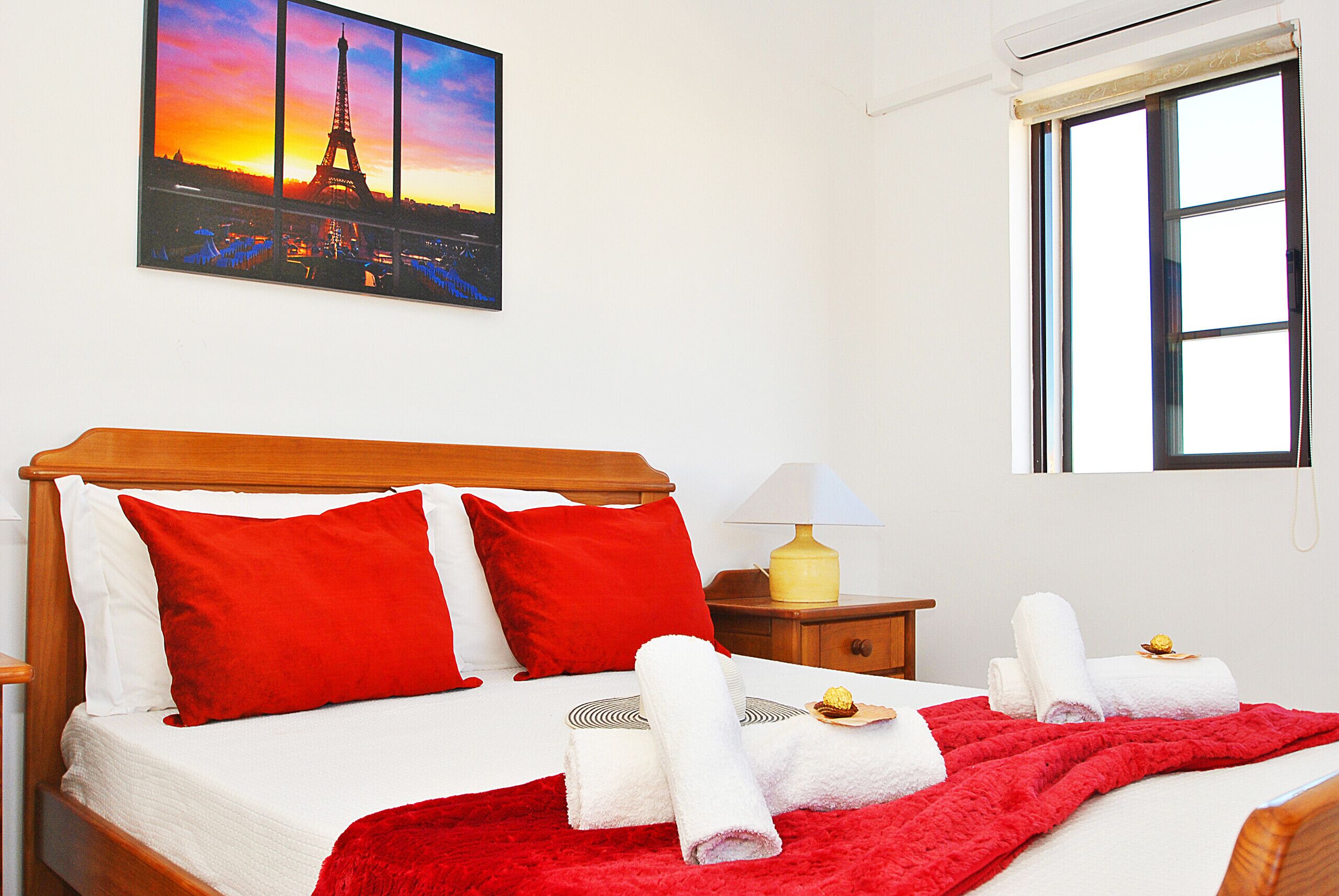 Una habitación de hotel con cama, almohadas rojas, manta roja, mesita de noche con lámpara, ventana y un cuadro de la Torre Eiffel.