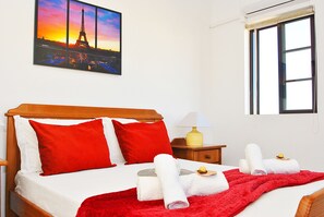A hotel room with a bed, red pillows, a red blanket, a nightstand with a lamp, a window, and a painting of the Eiffel Tower.