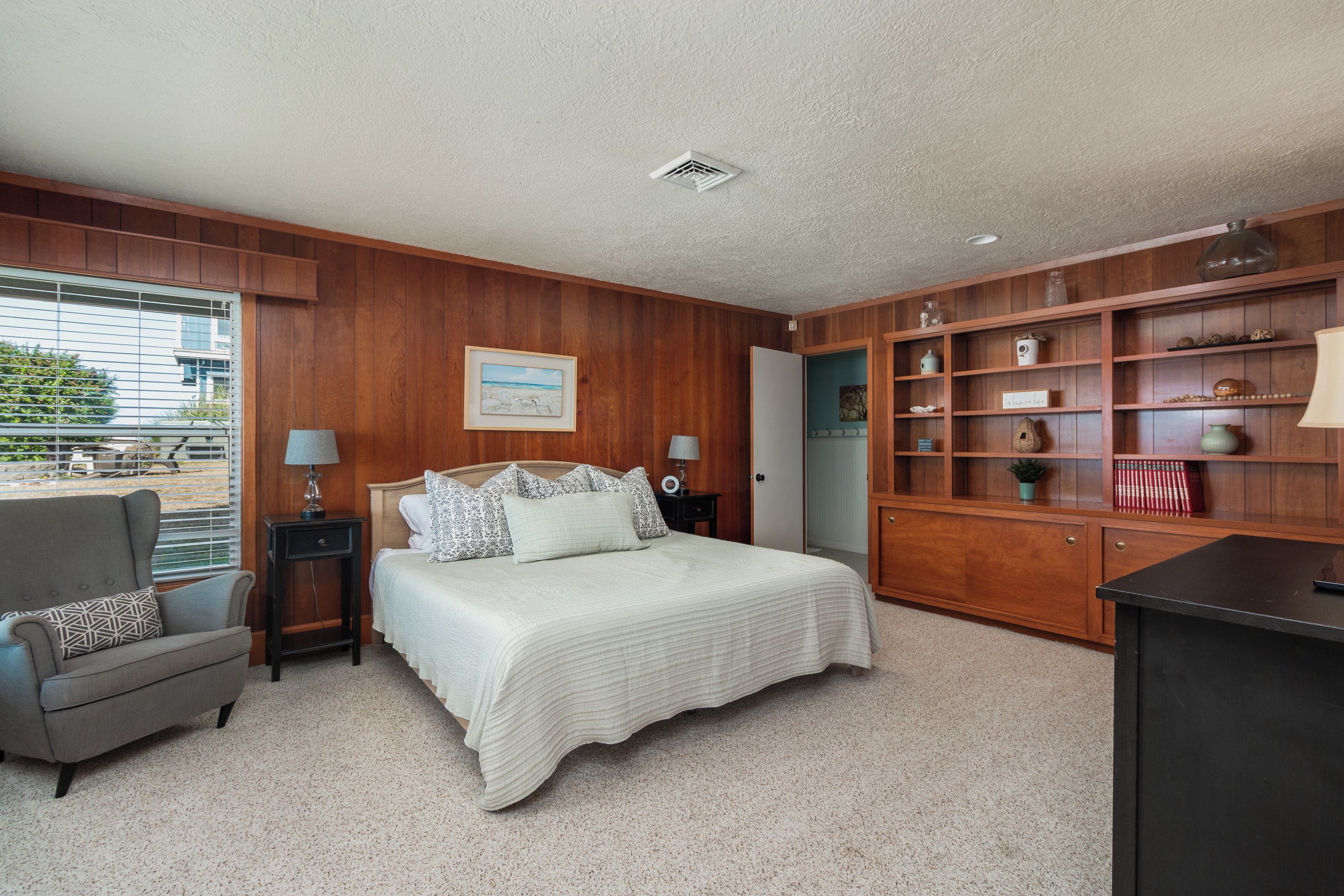 A bedroom with a bed, bedside tables, a chair, a bookshelf, and a window with blinds.