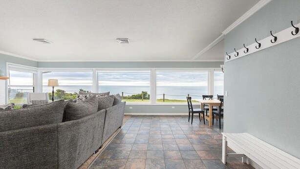 A room with a grey sofa, a dining table with chairs, and a view of the sea through large windows. - Oceanfront Home with Sweeping Pacific Views (Lincoln City)