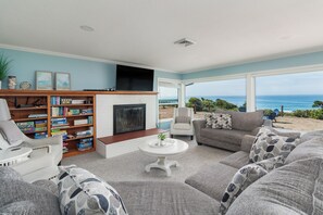Ein Wohnzimmer mit Kamin, Bücherregal, Fernseher und Meerblick.
