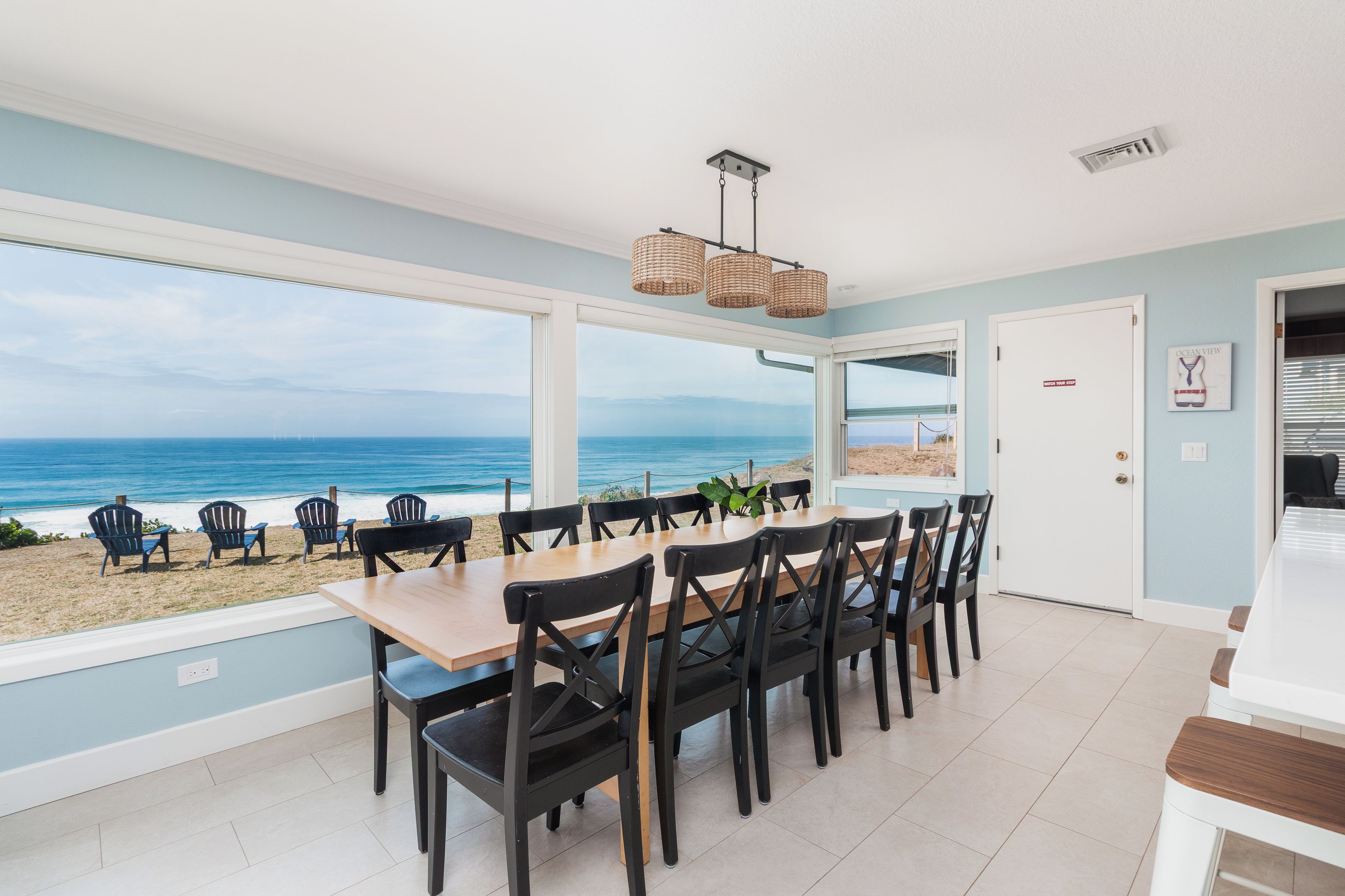 A dining area with a view of the beach and ocean, featuring a large table and chairs, a hanging light fixture, and a door leading outside.