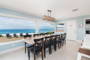 Ein Essbereich mit Blick auf Strand und Meer, ausgestattet mit einem großen Tisch, Stühlen, einer Pendelleuchte und einer Tür zum Außenbereich.