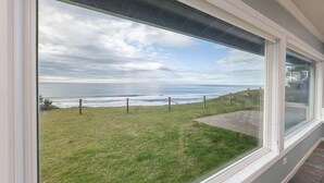 A view from a window overlooking a grassy area with a fence and a body of water in the distance. - Oceanfront Home with Sweeping Pacific Views (Lincoln City)