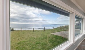 A view from a window overlooking a grassy area with a fence and a body of water in the distance. - Oceanfront Home with Sweeping Pacific Views (Lincoln City)