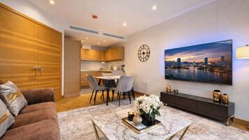A modern living room with a sofa, a coffee table with a vase of flowers, a dining area with chairs, a kitchen in the background, and a large cityscape painting on the wall.