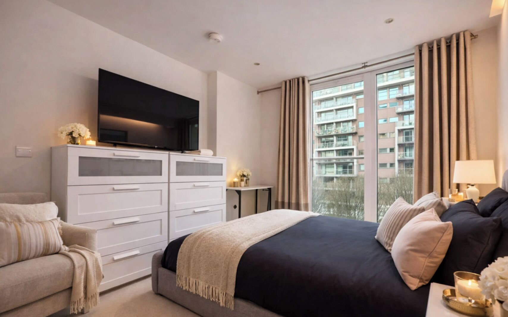 A hotel room with a bed, a TV, a desk, and a view of a building.