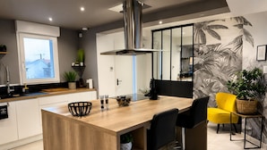 A modern kitchen with a central island, bar stools, and a dining area with a yellow chair. - Must-See / Jacuzzi - Pool - Sauna - Turkish Bath (Challans)