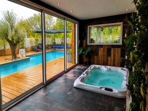 A modern indoor pool area with a hot tub, wooden deck, and large windows offering a view of the outdoors.