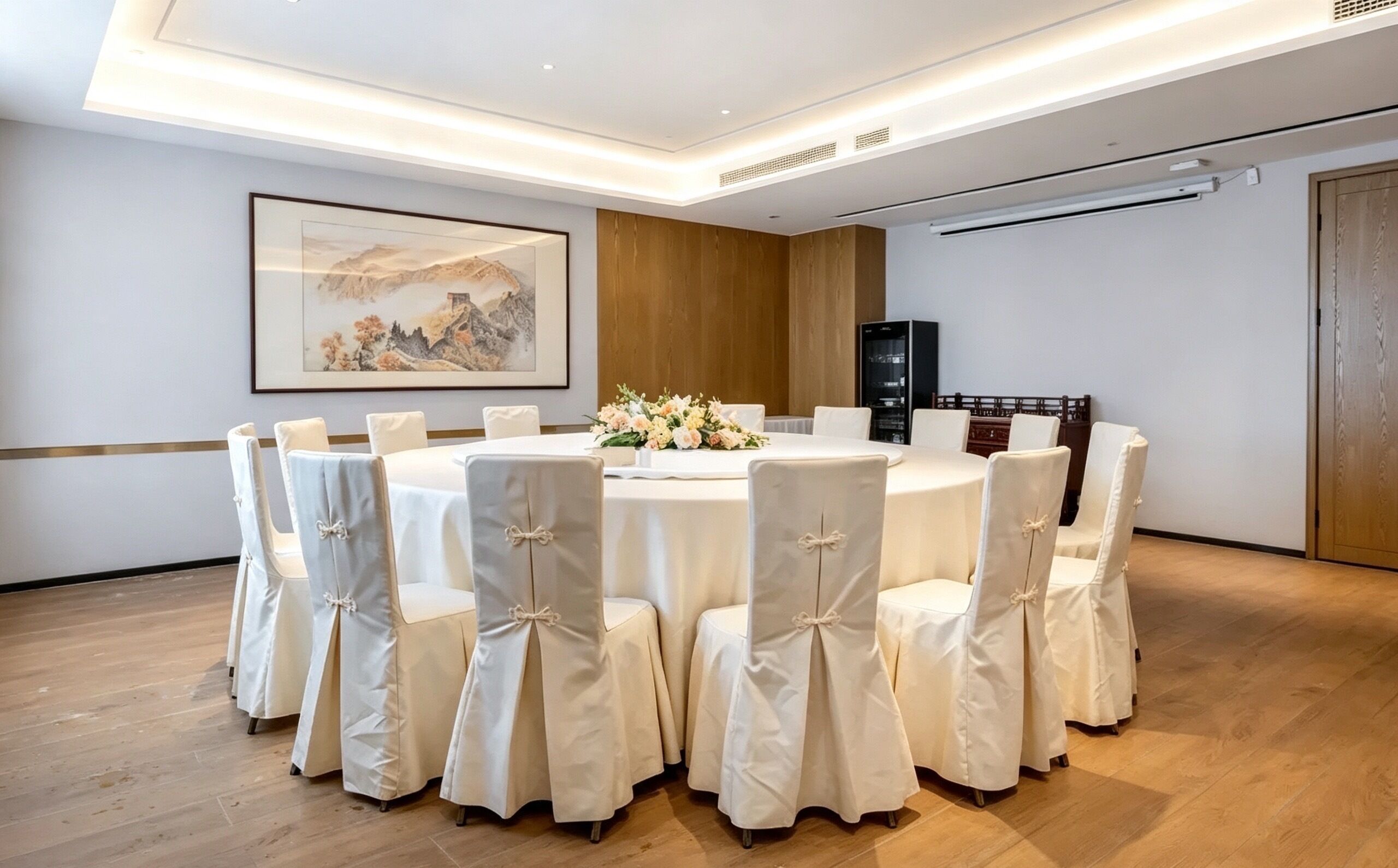 A conference room with a round table, white chairs with bows, a floral centerpiece, and a framed painting on the wall.