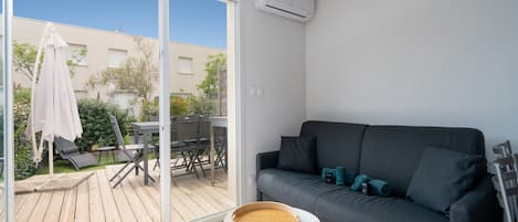 A modern living room with a grey sofa, a small round table with a tray, and a sliding glass door leading to an outdoor patio area.
