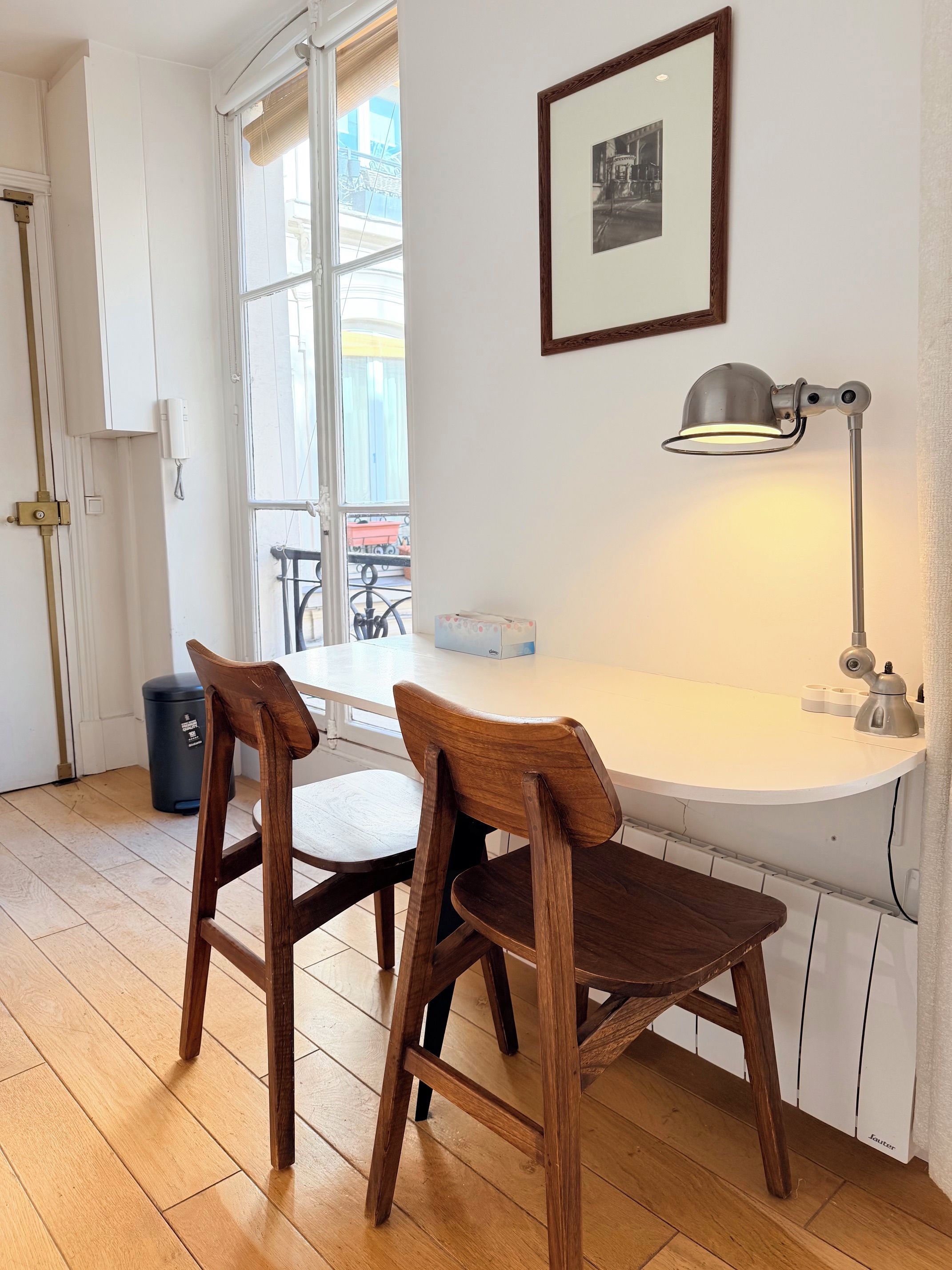 Ein Zimmer mit einem weißen Tisch, zwei Holzsesseln, einem gerahmten Bild an der Wand und einem Fenster mit Jalousien.
