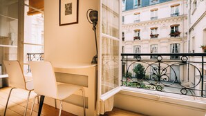 Ein Balkon mit weißem Stuhl, kleinem Tisch und einem Fenster mit Blick auf ein Gebäude mit Balkonen und Blumen.