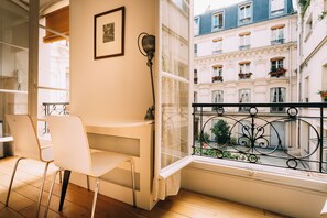 A balcony with a white chair, a small table, and a window with a view of a building with balconies and flowers.