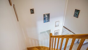 Una escalera de madera con barandilla, que lleva a un rellano con una pared blanca y una pared de piedra al fondo.