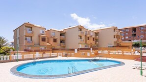 A swimming pool with a tiled deck, surrounded by a white fence, and adjacent to a multi-story building with balconies. - Caletillas BlackSand—ocean views—Wi-Fi—garage—2 bedrooms, pool. (Candelaria)