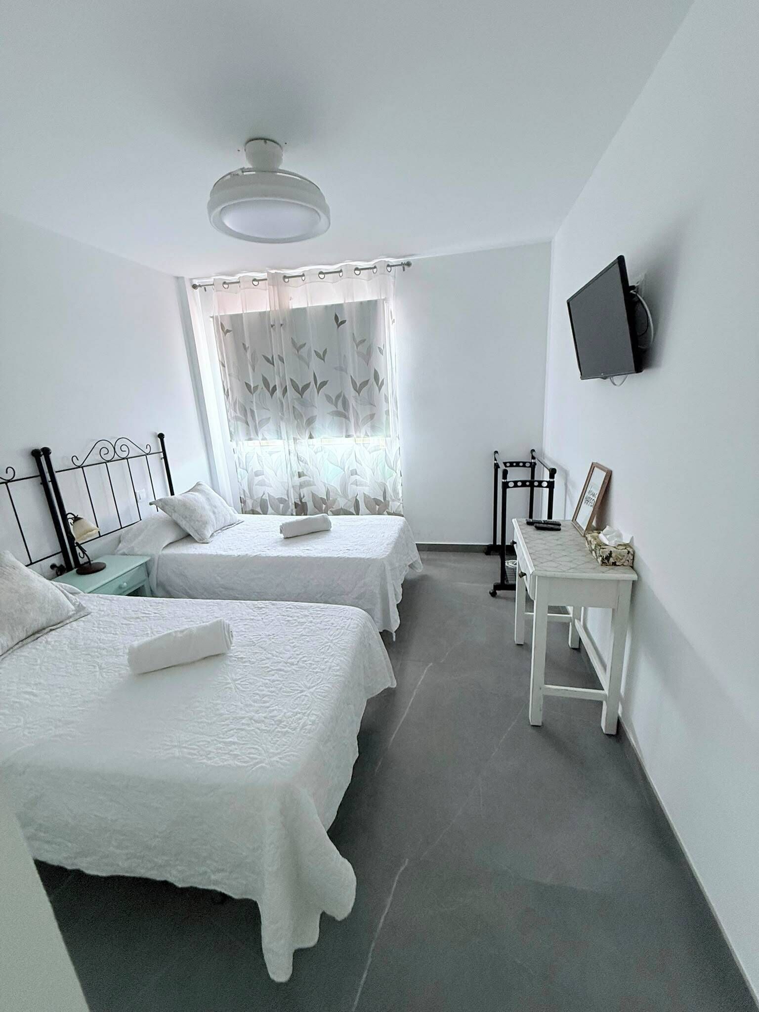 A room with two beds, a small table, a television on the wall, and a window with curtains.