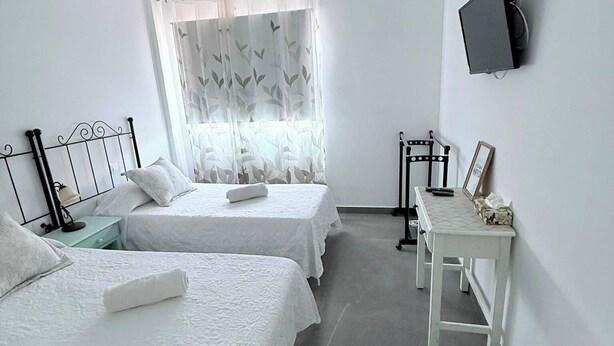 A room with two beds, a small table, a television on the wall, and a window with curtains. - Caletillas BlackSand—ocean views—Wi-Fi—garage—2 bedrooms, pool. (Candelaria)