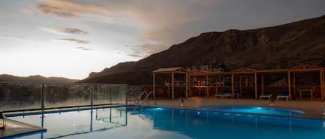 Una piscina con cerramiento de vidrio, rodeada por una terraza con sillas y mesas, con montañas al fondo.