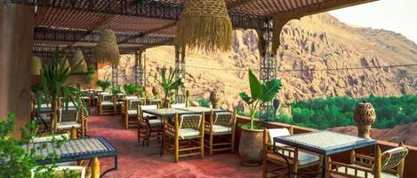 An outdoor dining area with tables and chairs, surrounded by plants and a mountainous backdrop.